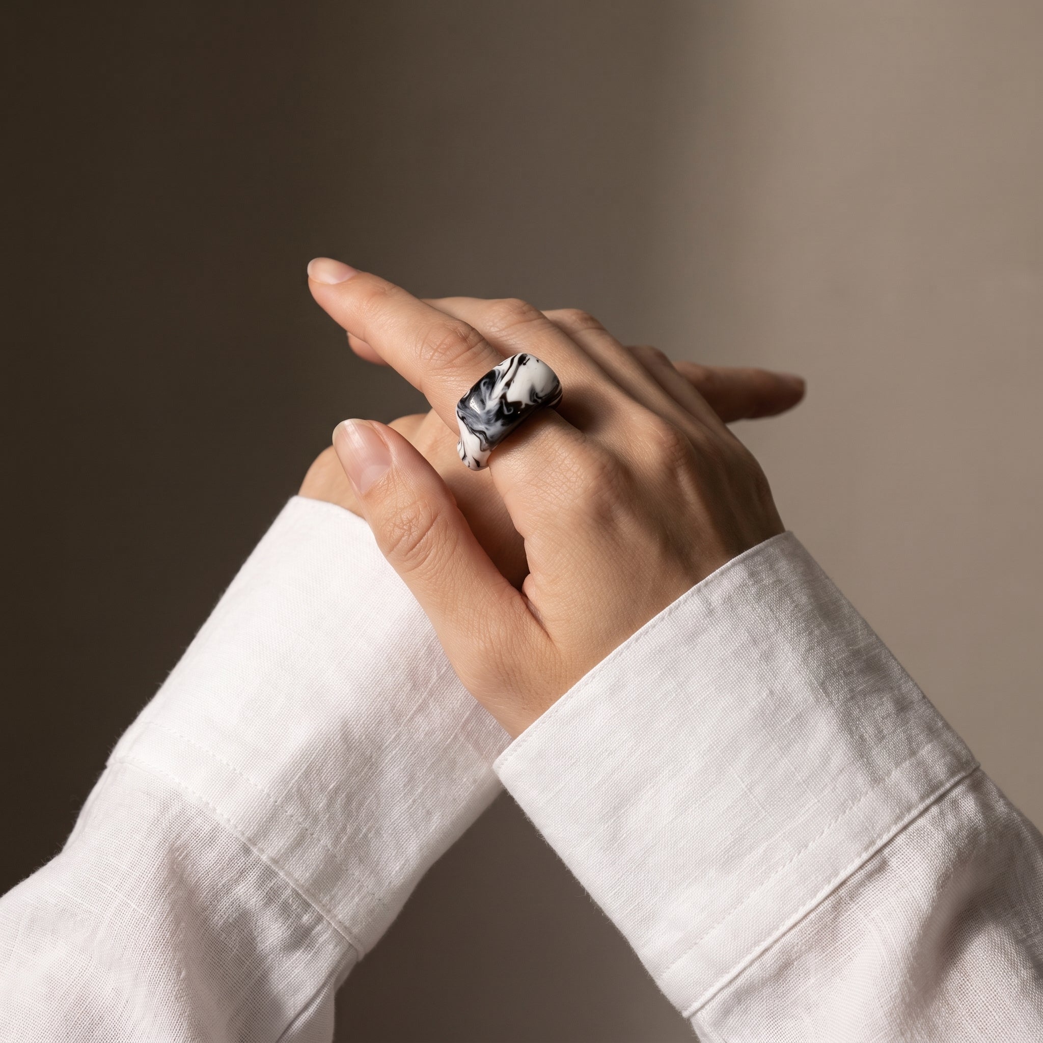 Petra Surco Black — anillo de porcelana negro con vetas blancas marmoladas en mano con camisa blanca lino, fondo tierra oscuro
