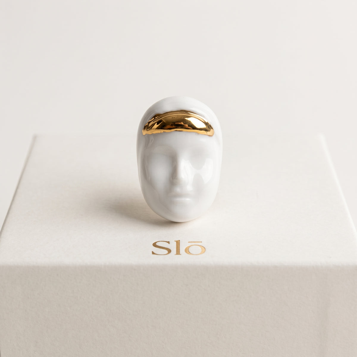 Domina — vista frontal del anillo de porcelana blanca con rostro y diadema dorada, presentado sobre caja de regalo Slō, fondo crema
