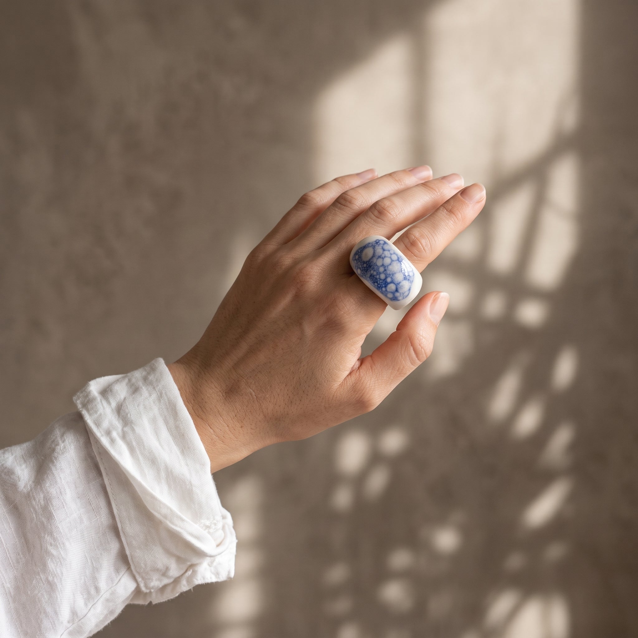Espuma de Mar — anillo de porcelana blanca con esmalte azul y burbujas en mano con camisa lino blanca, luz de ventana lateral sobre fondo tierra