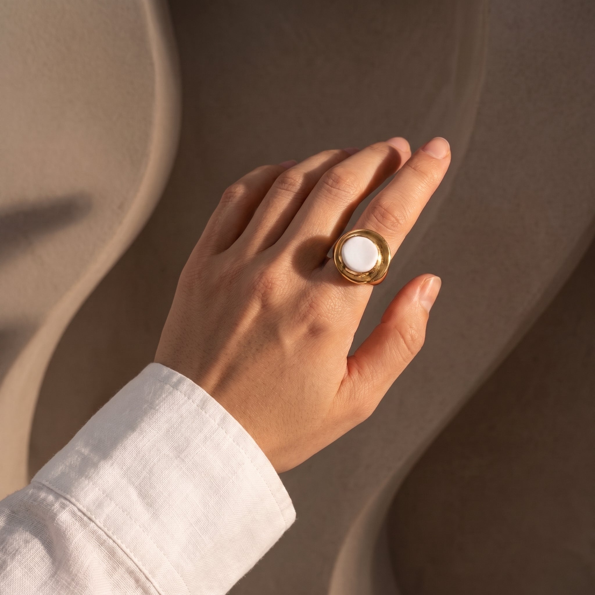 Eclipse Parcial — anillo de porcelana blanca con aro de oro ajustable en mano apoyada sobre superficie curva beige, luz solar lateral con sombras