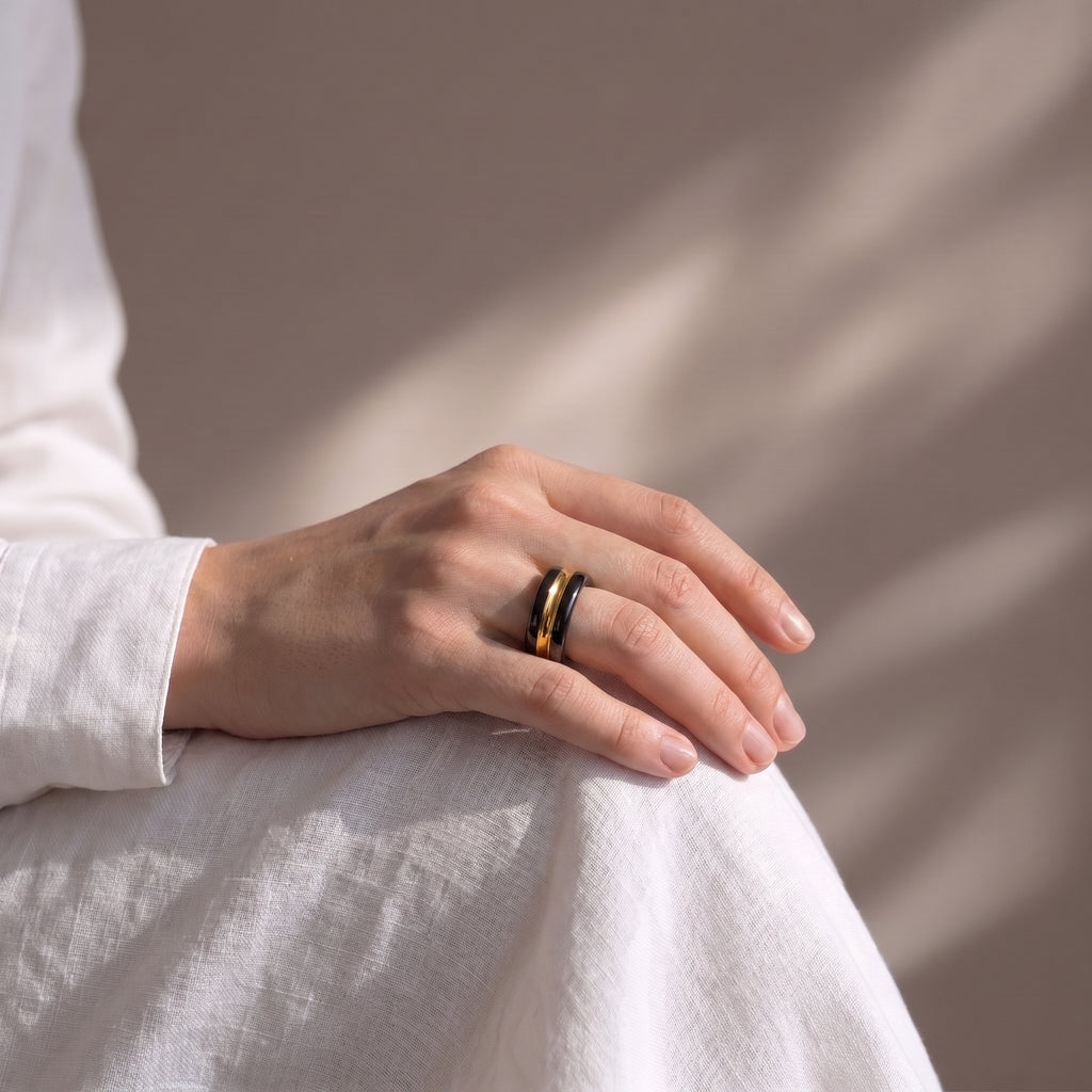 Trinity — anillo de porcelana negra con franja de oro central en mano apoyada sobre tela lino blanca, luz de ventana lateral con sombras suaves