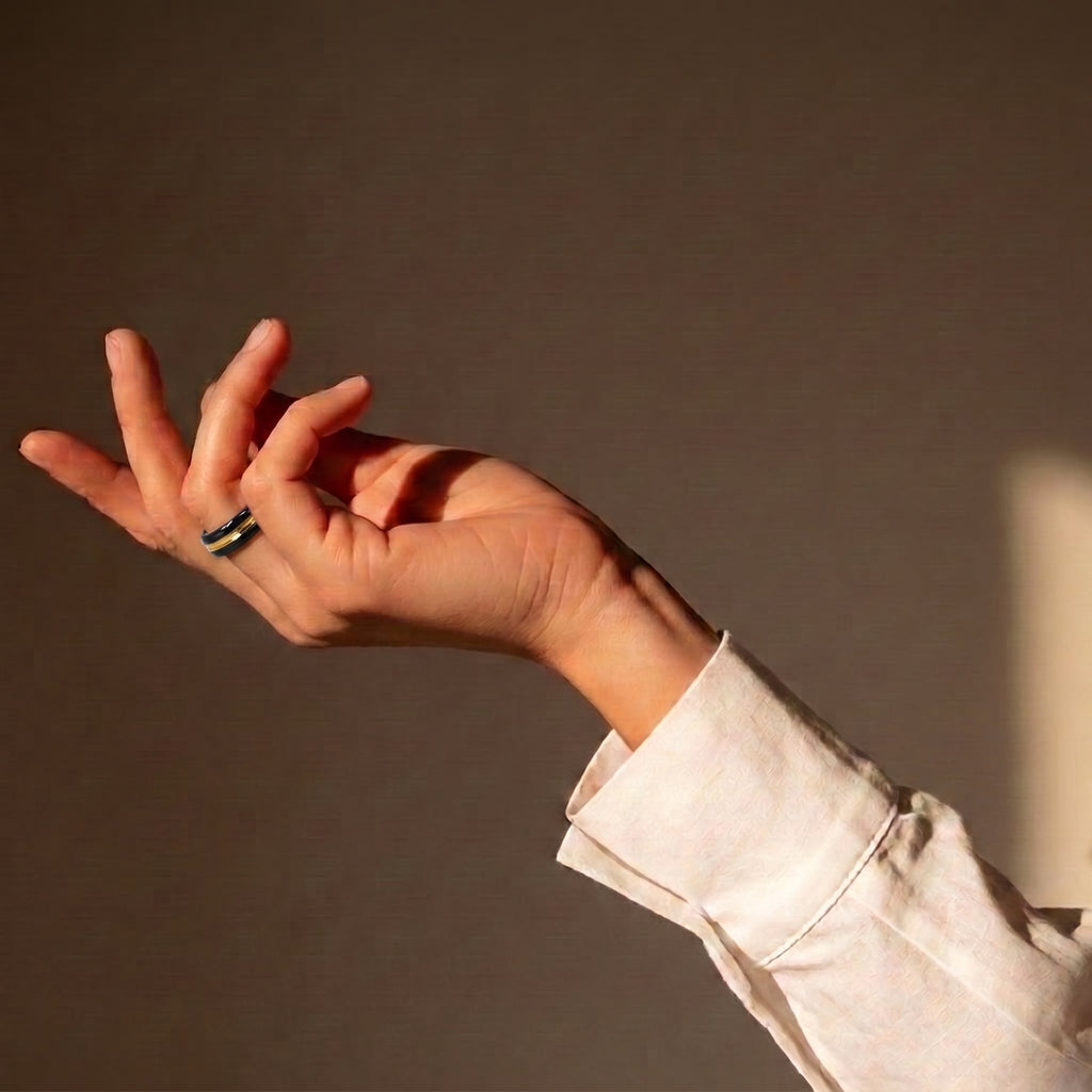 Trinity — anillo de porcelana negra con franja de oro central en mano alzada con camisa lino blanca, luz solar cálida sobre fondo tierra oscuro