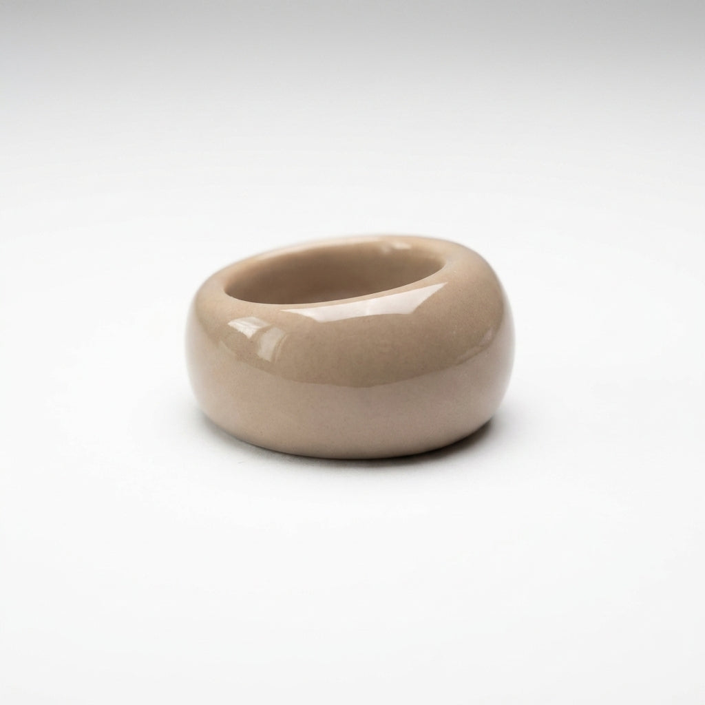 Dovela — anillo de porcelana con aro ancho y redondeado en esmalte topo brillante, vista en ángulo sobre fondo blanco mostrando el volumen y la forma de donut inflado
