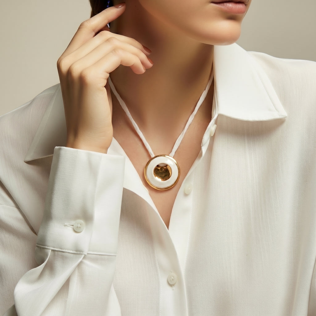 Eclipse Total colgante — disco de porcelana blanca mate con agujero central enmarcado por aro de oro brillante en cuello de mujer con camisa blanca, fondo beige con luz suave