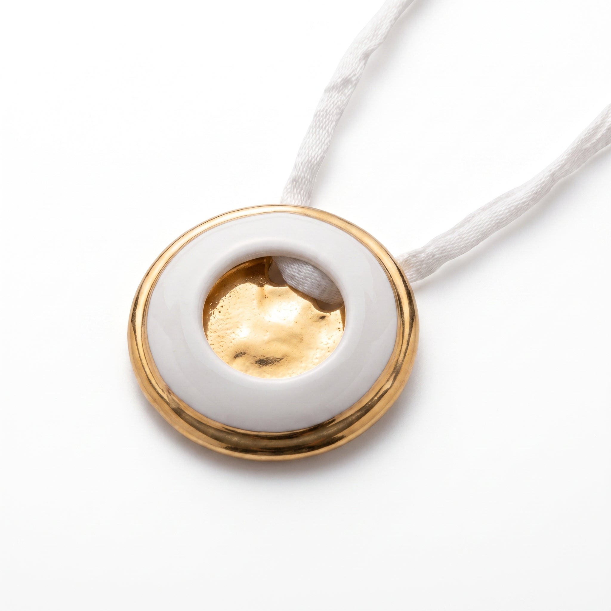 Eclipse Total colgante — disco de porcelana blanca con agujero central y aro de oro, vista en ángulo sobre fondo blanco con cordón de seda blanca trenzada mostrando el volumen y el acabado dorado