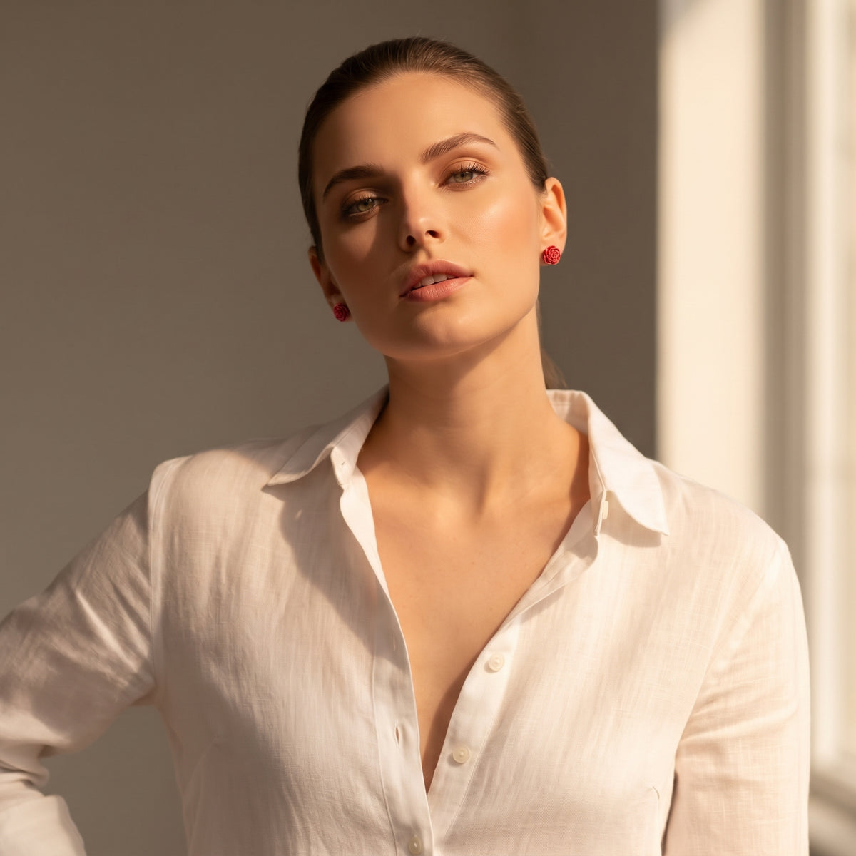 Pitimini Coral — pendientes de botón de porcelana coral puestos en modelo con camisa blanca y moño, luz dorada lateral que realza el color coral sobre piel