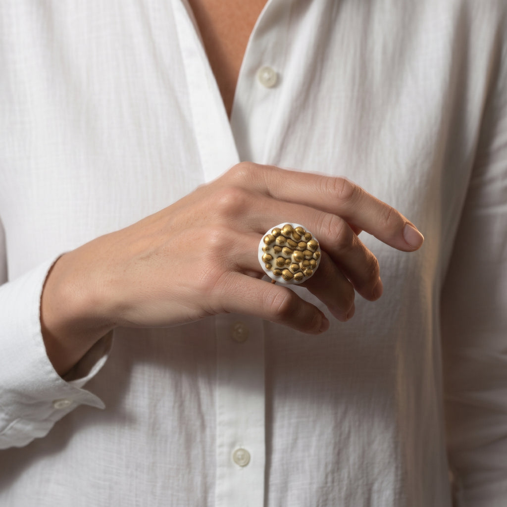 Maní Dorado — anillo de porcelana blanca con esferas doradas en dedo corazón de mano con camisa blanca, vista editorial mostrando la escala y la textura orgánica