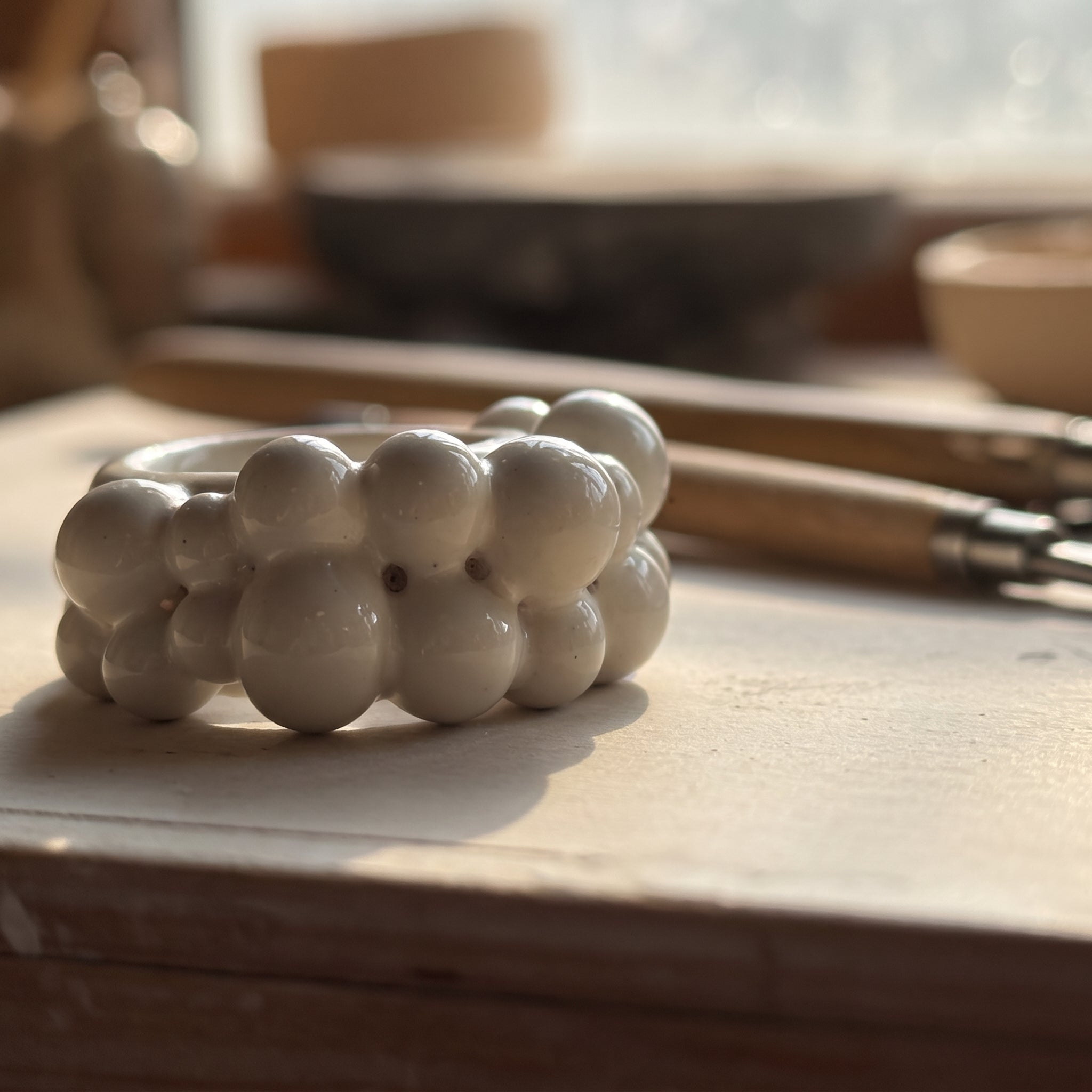 Burbujas — anillo de porcelana blanca con doble fila de esferas sobre mesa de madera con luz cálida de taller