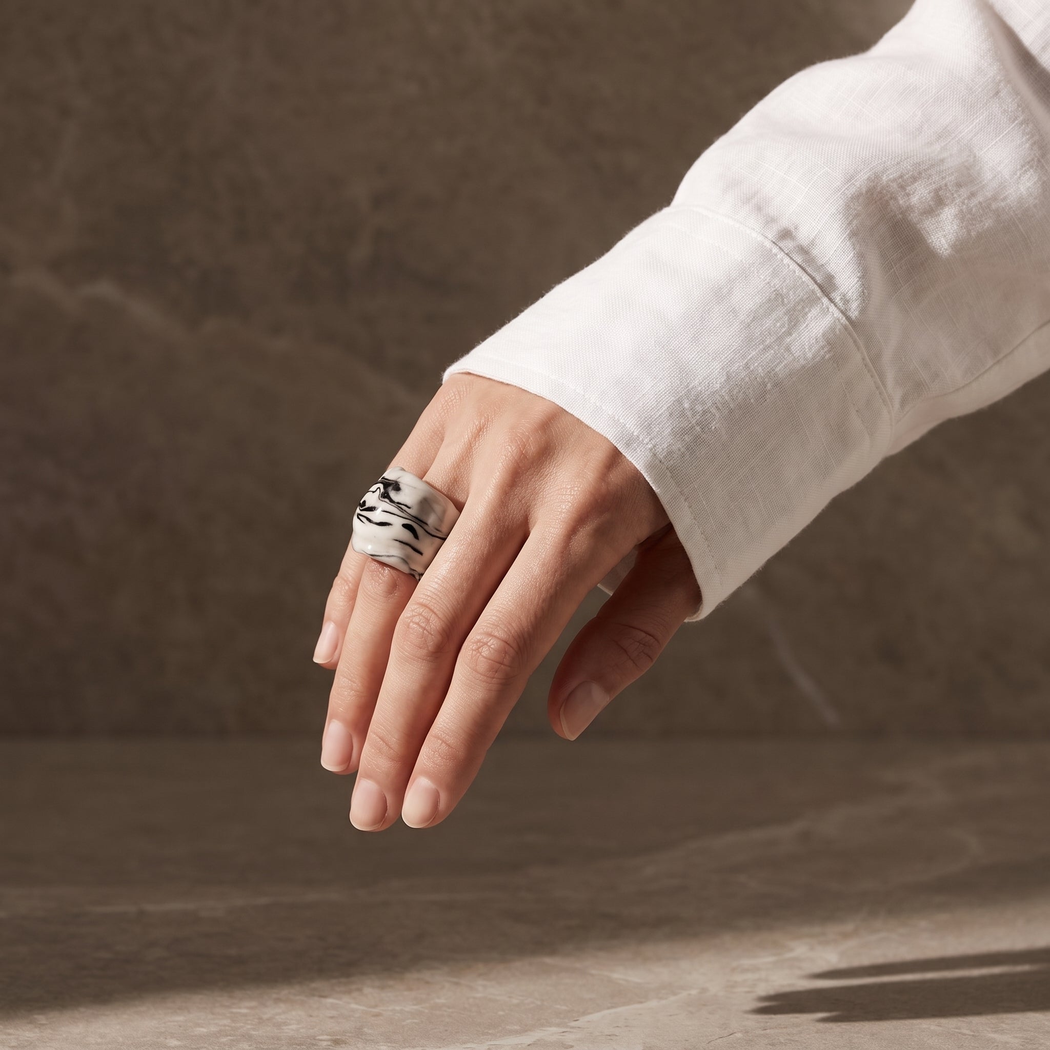 Petra Black — anillo de porcelana blanca con vetas negro intenso marmoladas a mano en mano caída con camisa blanca, fondo tierra oscuro