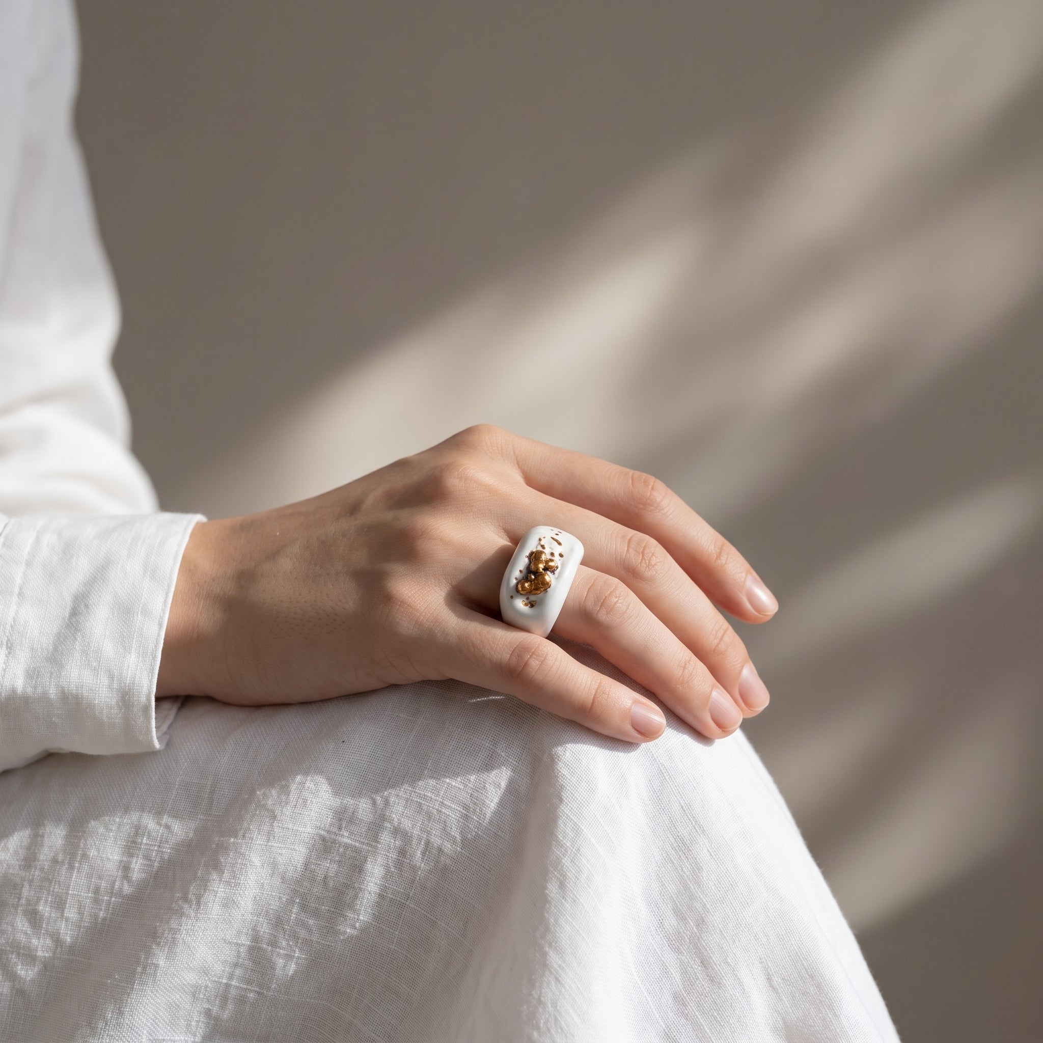 Navelgas — anillo de porcelana blanca con pepitas de oro irregulares en mano apoyada sobre tela lino blanca, luz de ventana lateral sobre fondo tierra