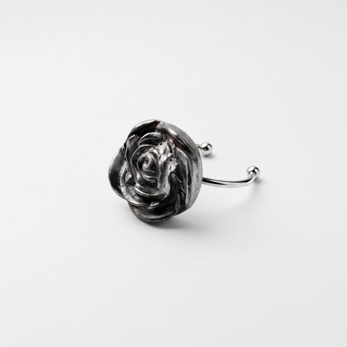 Camelia — anillo de porcelana platino en dedo índice, mano con camisa blanca sobre fondo beige cálido mostrando la escala y presencia de la flor