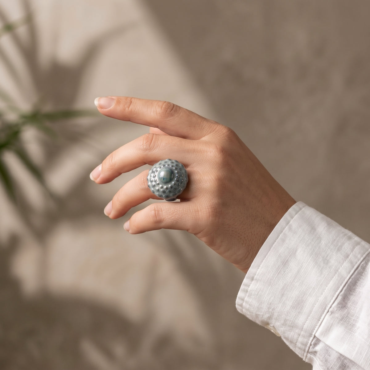 Girándola — anillo de porcelana en acabado verde celadón con esfera de bultos y aro fino dorado, vista en mano con camisa blanca sobre fondo beige con sombra de planta