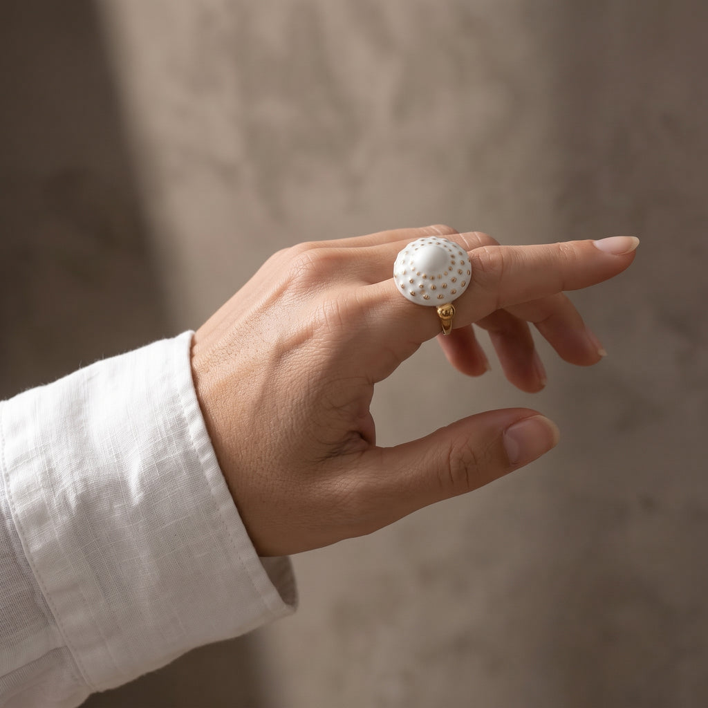 Girándola Blanco Oro S — mano apoyada en borde de mármol gris con anillo de porcelana blanca y puntos dorados, camisa blanca, ventana al fondo