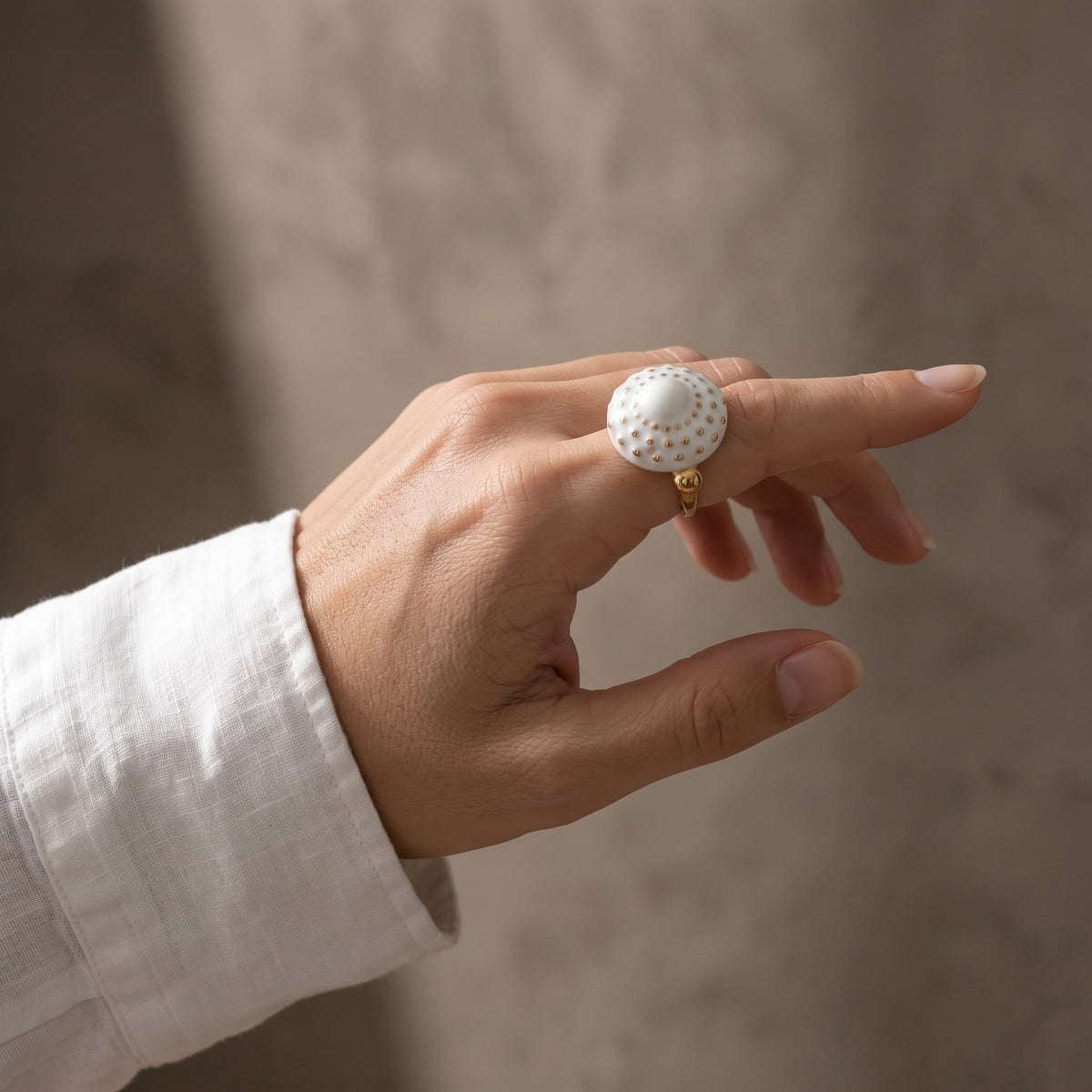 Girándola Blanco Oro S — mano apoyada en borde de mármol gris con anillo de porcelana blanca y puntos dorados, camisa blanca, ventana al fondo